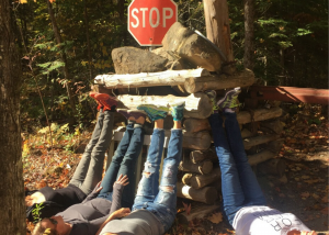 A photo of women laying on the ground, with their feet resting on a pile of logs and rocks, with a stop sign behind the pile.